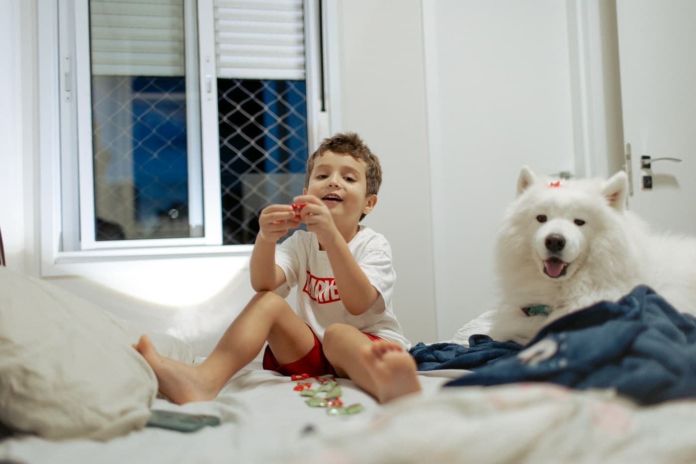 Menino sorrindo sentado na cama ao lado do seu cachorro branco